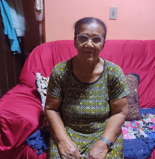 Senhora sentada em sofá vermelho, com roupas tradicionais, em casa simples e acolhedora