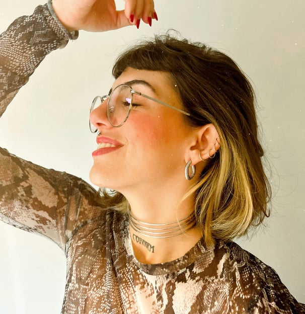 Foto de mulher sorrindo com óculos de grau piercings na orelha e boca, brincos e chocker no pescoço.