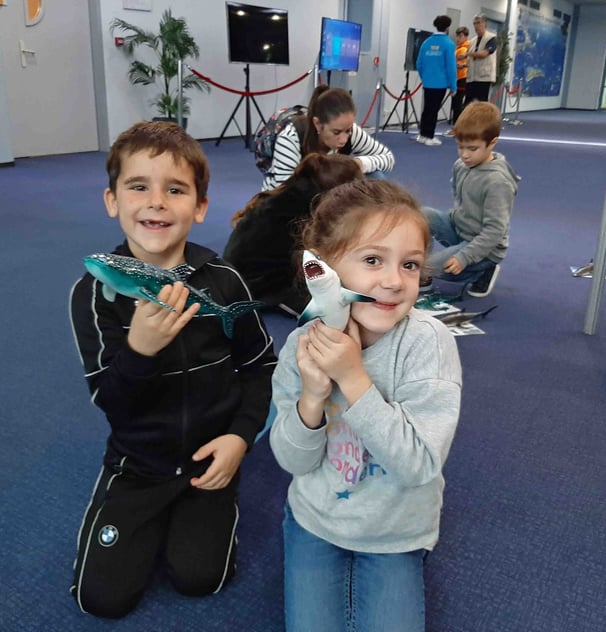 2 enfants à genoux souriants qui tiennent des figurines de requins dans les mains