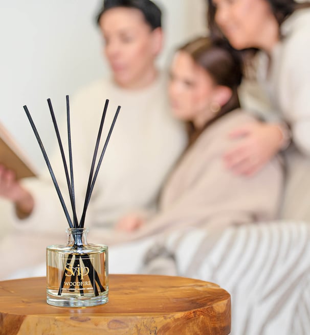 a group of women sitting on a couch reading a book and they sit behind a bottle of home scent