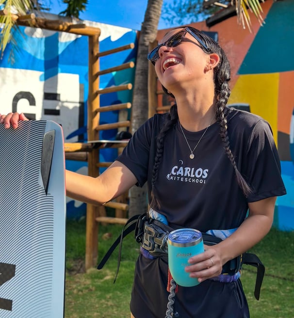 mulher jovem, sorrindo segurando um copo e uma prancha de kitesurf