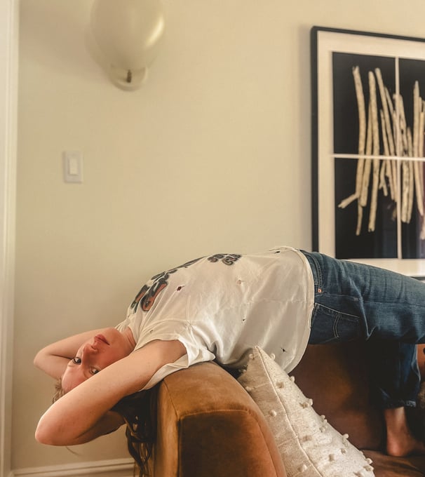 Woman reclining upside down on a copper sofa in a cozy room with modern art, wearing casual clothes and appearing relaxed.