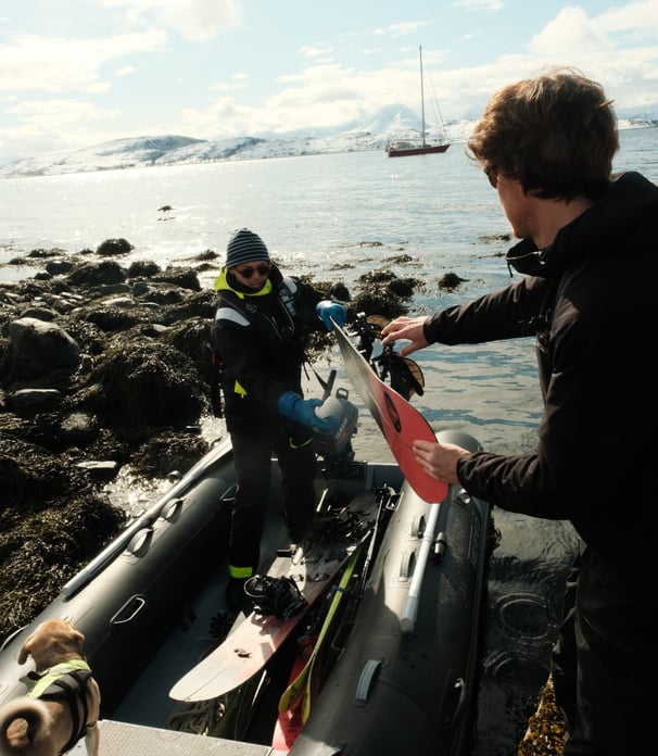 Two skiers unloading gear from a dinghy with snowy Lyngen Alps and a sailboat in the background