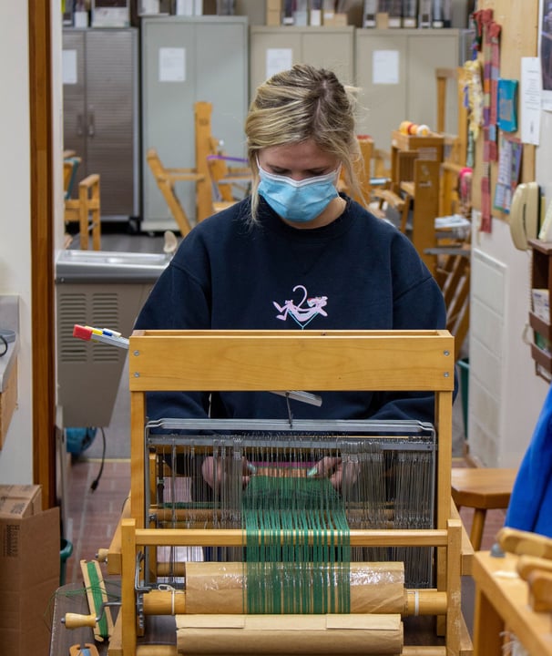 A young woman standing in front of a table loom adjusting your piece.