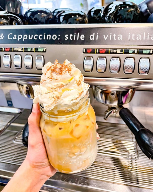 a person holding a jar of ice latte with whip cream by espresso machine