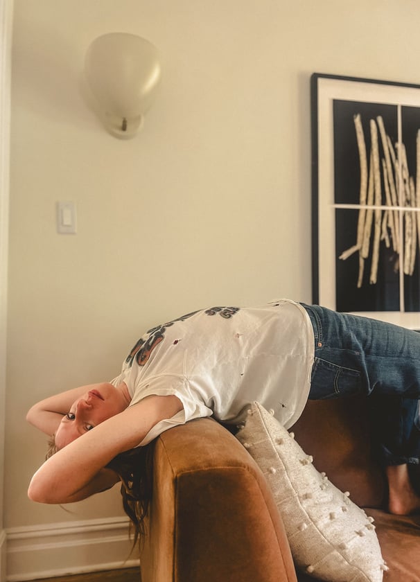 Woman reclining upside down on a copper sofa in a cozy room with modern art, wearing casual clothes and appearing relaxed.