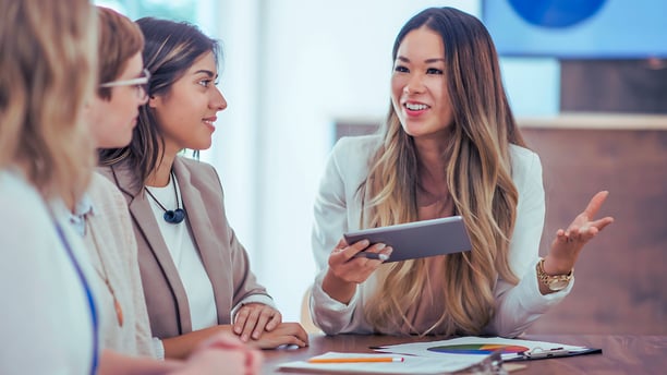 Asian business consultant discusses data while holding a tablet in front of three other female leaders in a boardroom.