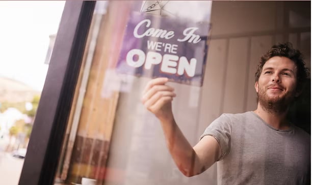 Man turning around Come In We're Open sign on coffee shop window
