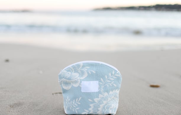Trousse de maquillage ÉTOFÉ en tissu recyclé bleu clair à motifs floraux, posée sur une plage