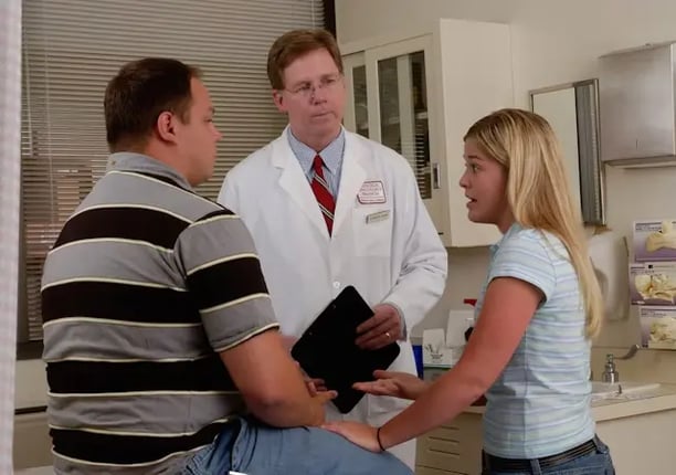 A doctor converses with a man and woman in a hospital setting, discussing medical concerns.