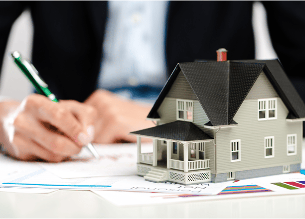 A miniature model of a house with a black roof and grey walls sits on a table with documents and cha