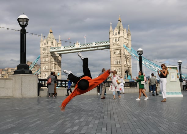 YoYo breakdancing with Tower Bridge, London, in the background as passers-by watch.