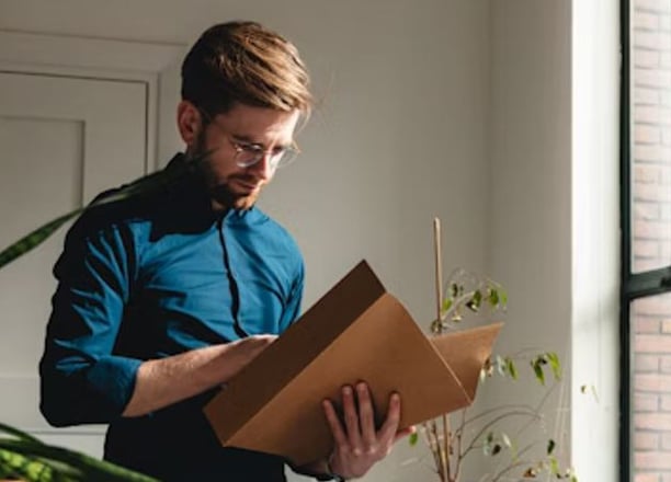 Bookkeeper standing up in a blue shirt looking at a client's folder to analyze their finances. A plant is to their left.