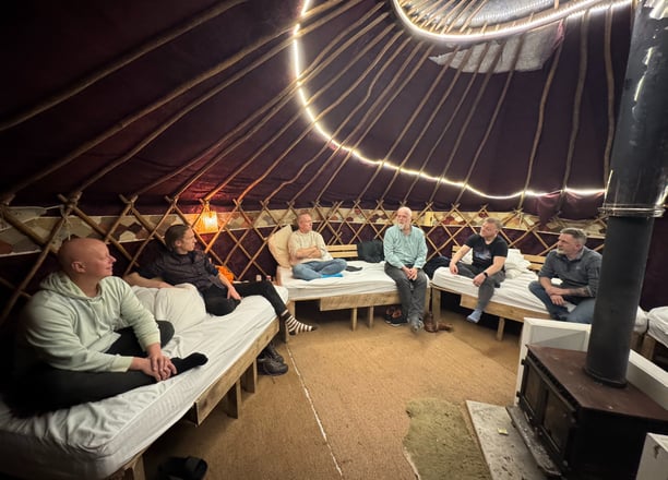 Photo of mens group with six men taking place in a yurt with wood stove