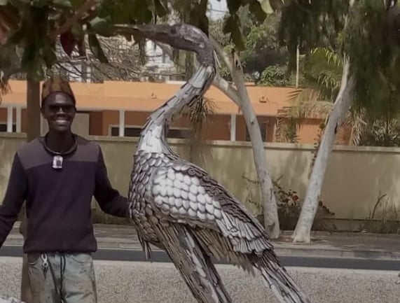Moussa Fofana posant avec une sculpture métallique d’un héron géant fabriqué à Dakar