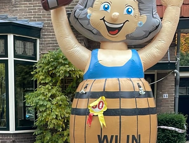 Opblaas pop Sarah met blauwe jurk in bruin wijnvat aan de voorkant van een huis