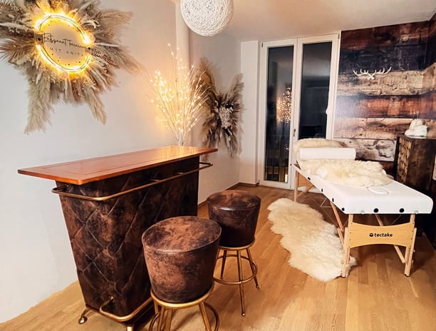 Auf dem Bild ist eine braune Bar, mit zwei braunen Barhockern, Massageliege mit weißem Leder, auf der ein weißes Lammfell