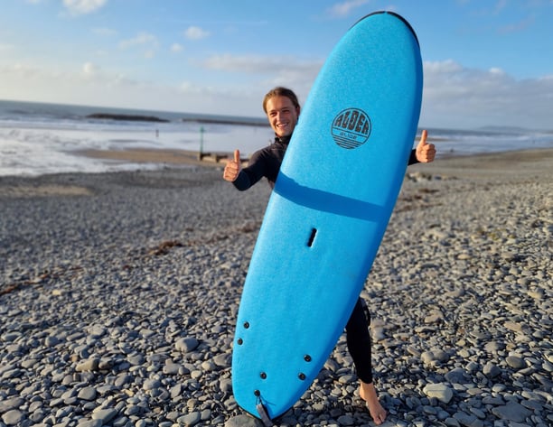 Surf coach Celynholding a blue surfboard and giving a double thumbs up