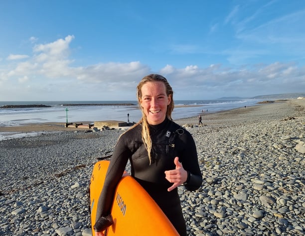 Surf coach Cerys holding an orange surfboard and giving a shaka