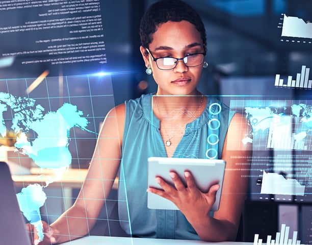 A female leader with glasses holds a tablet and types on a laptop at her desk, surrounded by holographic maps and graphs.