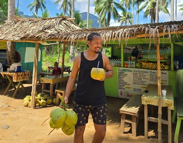 a man holding a drink in front of a hut with coconut trees