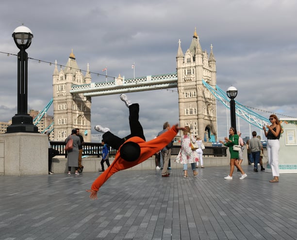 YoYo breakdancing with Tower Bridge, London, in the background as passers-by watch.