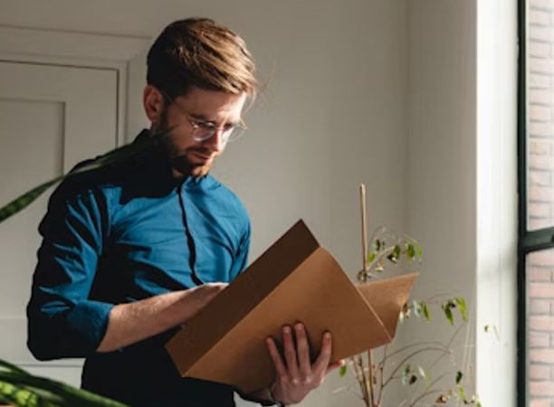 Bookkeeper standing up in a blue shirt looking at a client's folder to analyze their finances. A plant is to their left.