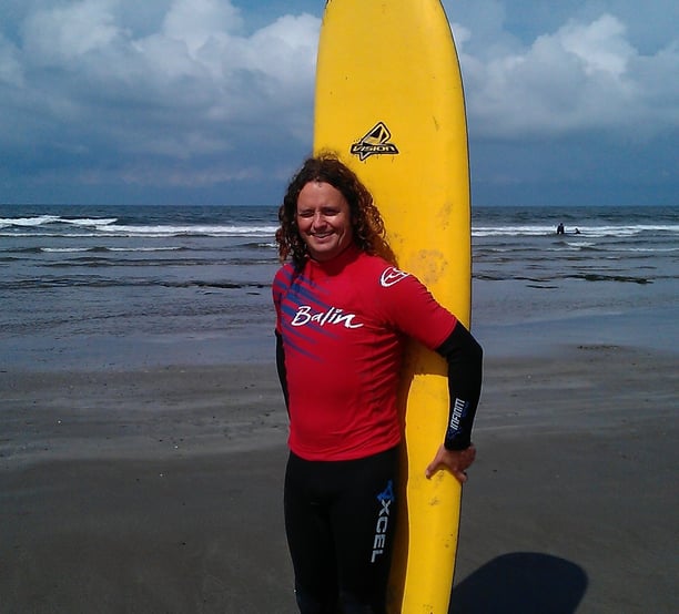 Surf school owner Gray satnding with a yellow surfboard behind him at the beach