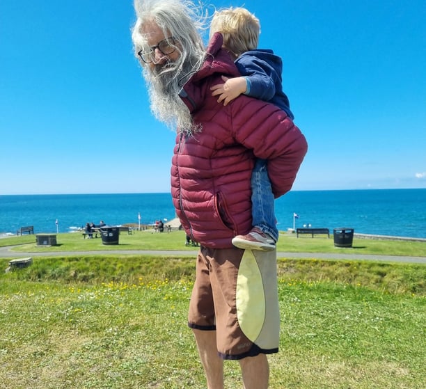 Head surf coach Simon stood with a little boy having a piggy back in green field by the sea.