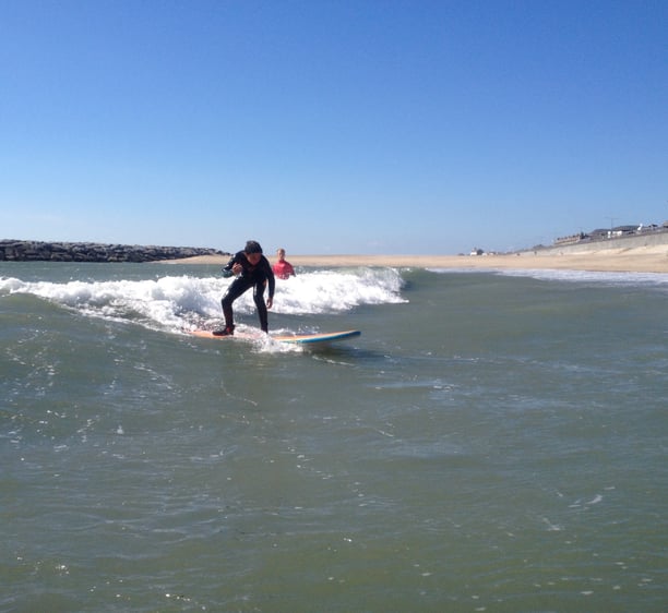 A young male surfer rides an unbroken wave with sunny skies while a surf instructor looks on