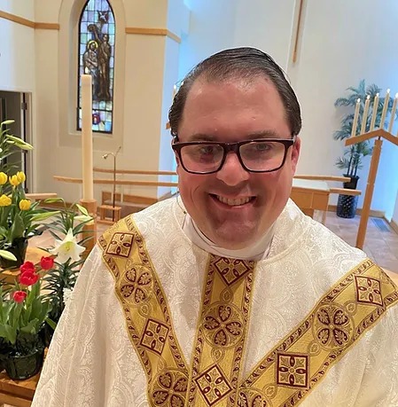 Person in religious vestments standing in a church with flowers and stained glass window in the back