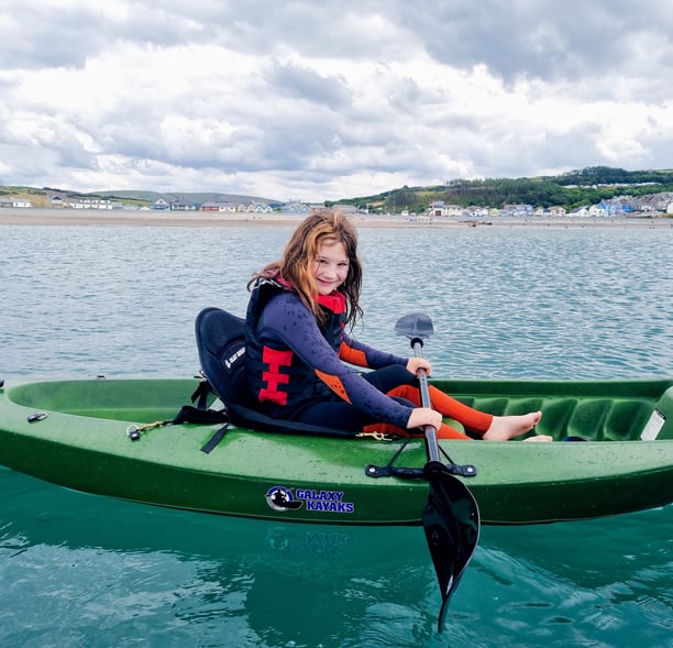 A girl kayaking in a sit on top kayak in the sea with Borth village and cloudy skies behind her 