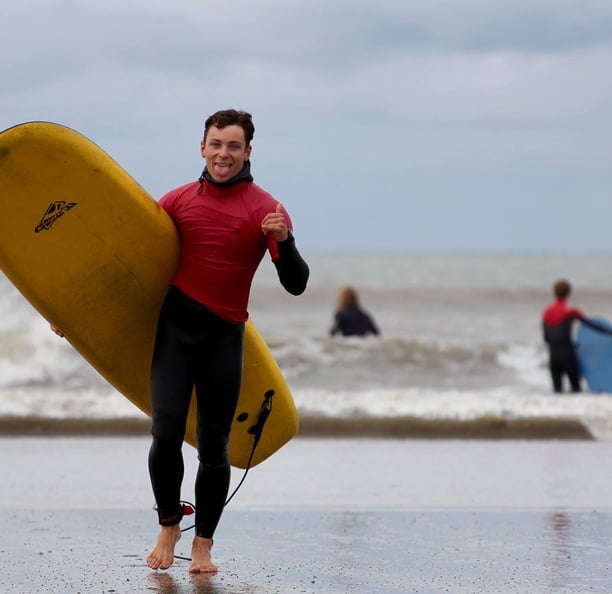 A stoked surf instructor carrying a yellow surfboard, giving a shaka and sticking his tounge out.