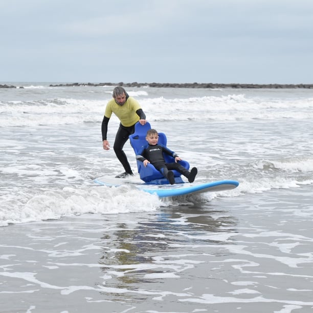 Young surfer in a seated surfboard with a coach controlling the board riding a wave