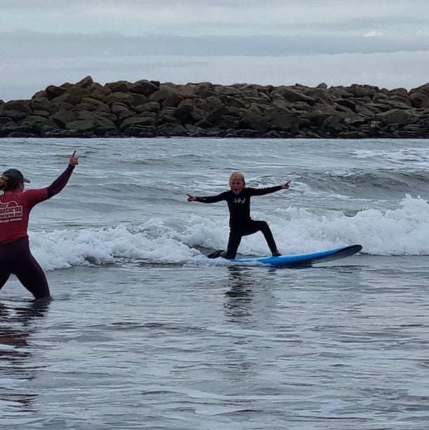 A young male surfer riding a wave cheerring as a female surf coach gives him encouragment.