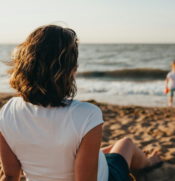 Femme assise seule sur la plage, face à la mer, et regarde un petit garçon jouer au bord de l'eau