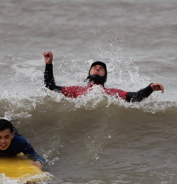 A prone smiling surfer dropping down a wave while a surf instructor jumps backwards over a wave.