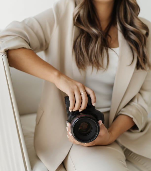 A woman in a beige jacket and white top, sitting on a chair holding a camera.