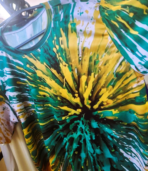 A imagem mostra uma camiseta impressa em sublimação, com uma explosão de tintas verde e amarelo