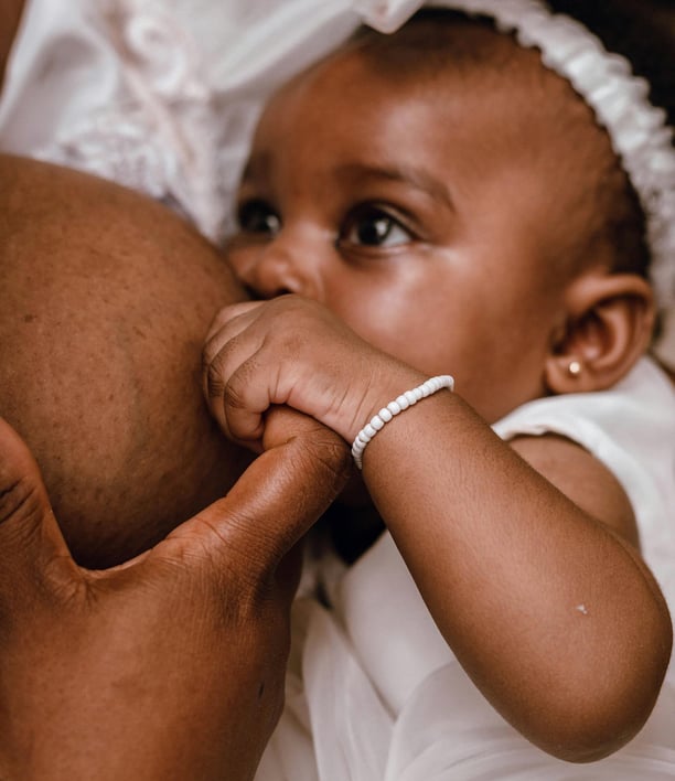 Bébé fille vêtue de blanc noire tétant le sein de sa maman et lui tenant le doigt