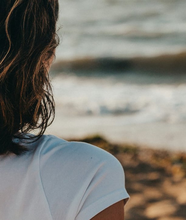 Femme seule vue de dos, assise sur la plage, face à la mer. Un enfant joue au loin, au bord de l'eau