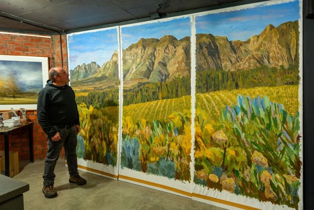 Martin Osner standing in his studio beside a large triptych landscape painting showing vineyards, fynbos, and mountains
