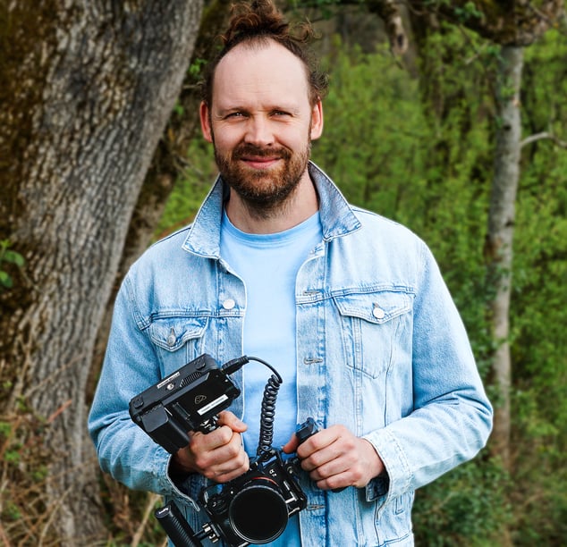 Lächelnder Mann mit Bart und Zopf in blauer Jeansjacke, draußen, Kamera Rig in den Händen, Porträt