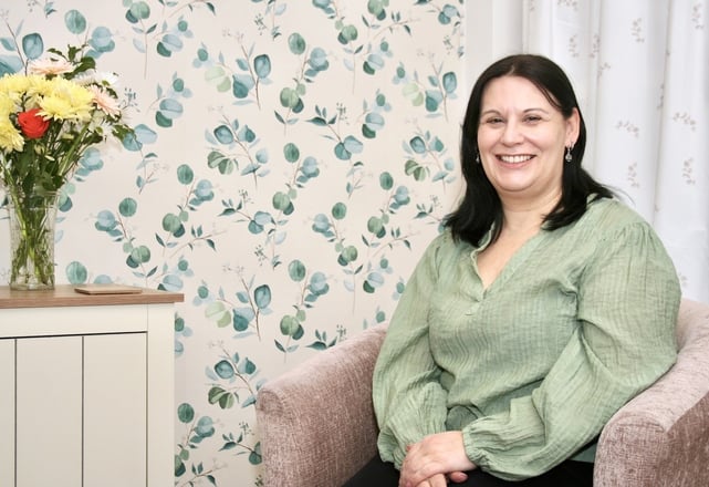 lady in green blouse sat in chair in front of eucalyptus wallpaper, next to sideboard with flowers