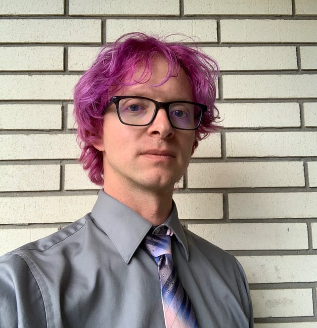 Tomkins with a grey shirt and blue and pink tie, with maroon colored hair and glasses.