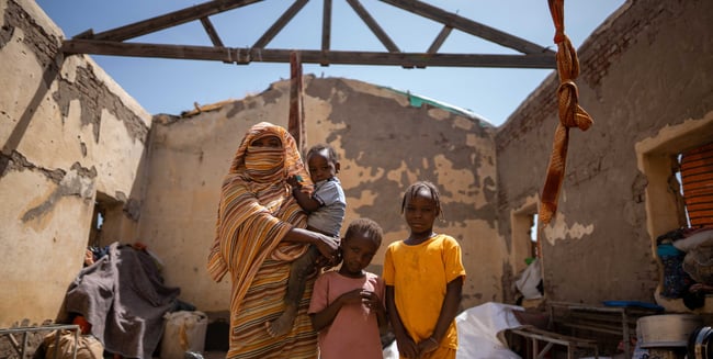 Sudanese woman and children standing inside a roofless home damaged by conflict.