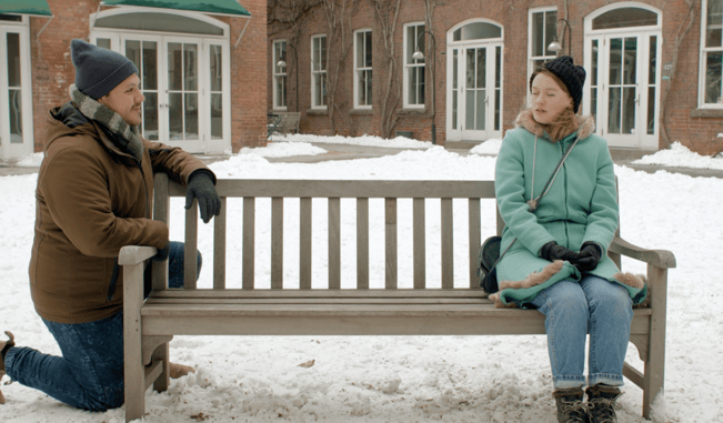 A young man leans on one end of a park bench, while a young woman sits eyes closed on the other end