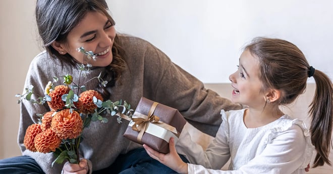Niña con camisa blanca dando regalo a mamá con sueter gris y sostentiendo flores