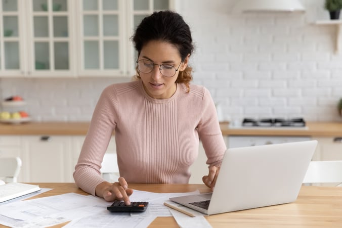 A woman sitting at her kitchen counter working on her calculator and laptop.