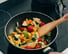 The image shows a stir-fry with bell peppers, tomatoes, and other vegetables being stirred in a pan.